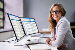 Woman wearing black-rimmed glasses and white blazer sitting in front of a double monitor with spreadsheets